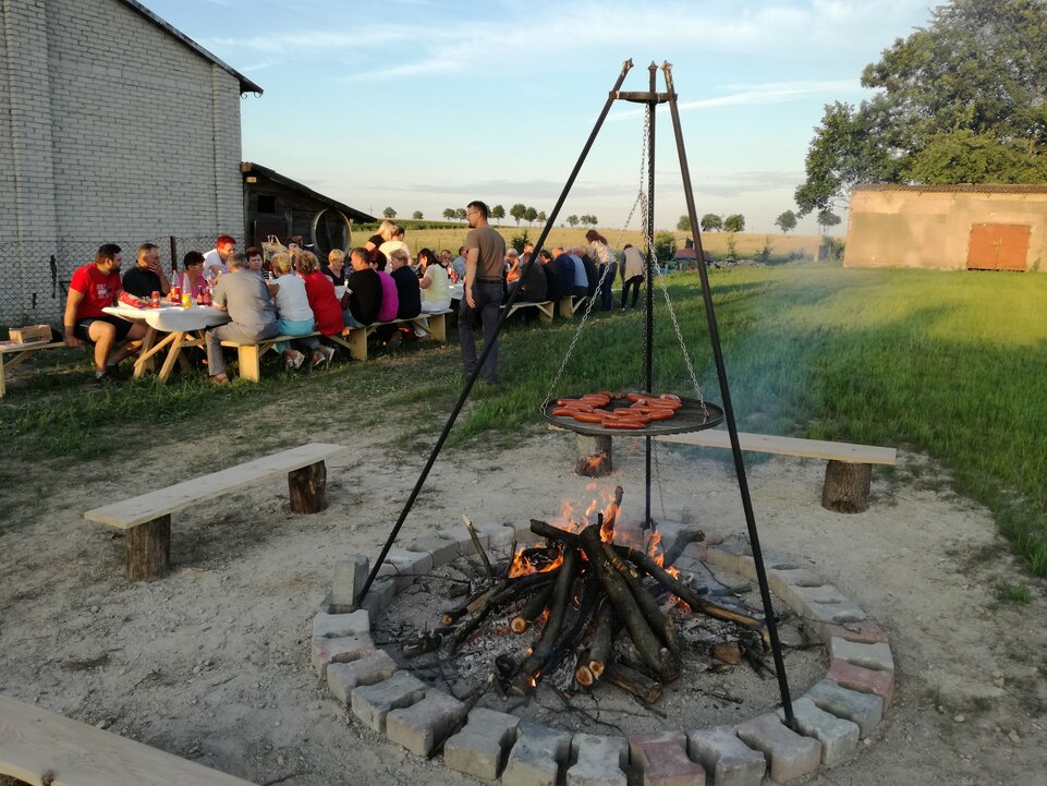 Impreza KGW Świniarów - na pierwszym planie ognisko z paleniskiem, na drugim uczestnicy imprezy siedzący za stołem